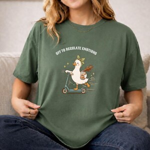 May include: A moss green short-sleeve t-shirt featuring a graphic of a white duck wearing a green headband, riding a blue scooter, and holding a brown beverage. The text above the duck reads, "OFF TO REGULATE EMOTIONS" in white capital letters.
