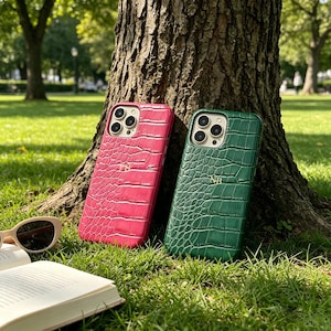 Può includere: Due custodie per telefono con texture a pelle di coccodrillo. Una è rosa acceso, l'altra verde scuro. Entrambe le custodie sono appoggiate a un tronco d'albero su un prato. Occhiali da sole e un libro aperto in primo piano.