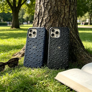 May include: Two phone cases, one navy blue and one black, with a textured pattern, are displayed on green grass next to a tree. The navy case has the initials "AX" and the black case has "TR". Sunglasses and a book are also visible.