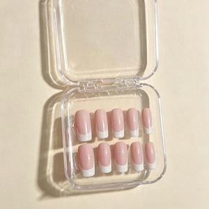 以下が含まれることがあります： ソフトピンクのベースと鮮やかなホワイトのチップが特徴の、クラシックなフレンチマニキュアデザインのネイルチップ10本セット。ネイルチップは、ヒンジ付きの蓋が付いた透明な正方形のプラスチック製収納ケースの中に、5本ずつ2列に並べられています。