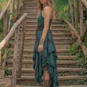 A woman stands on a rustic wooden staircase in a forest, wearing a forest green and black tie-dye fairy-style dress. The dress features a corset-style lace-up bodice and a high-low handkerchief hemline with layered pointed edges. She is styled with bohemian silver jewelry and fringe boots. The lighting is soft and natural, emphasizing the earthy, mystical, and witchy aesthetic of the outfit. Perfect for festivals, woodland ceremonies, or gothic boho fashion