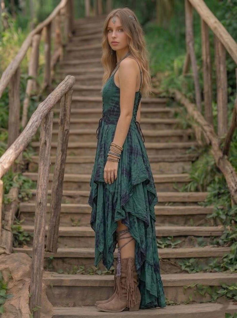 A woman stands on a rustic wooden staircase in a forest, wearing a forest green and black tie-dye fairy-style dress. The dress features a corset-style lace-up bodice and a high-low handkerchief hemline with layered pointed edges. She is styled with bohemian silver jewelry and fringe boots. The lighting is soft and natural, emphasizing the earthy, mystical, and witchy aesthetic of the outfit. Perfect for festivals, woodland ceremonies, or gothic boho fashion