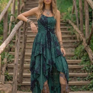 A woman stands on a rustic wooden staircase in a forest, wearing a forest green and black tie-dye fairy-style dress. The dress features a corset-style lace-up bodice and a high-low handkerchief hemline with layered pointed edges. She is styled with bohemian silver jewelry and fringe boots. The lighting is soft and natural, emphasizing the earthy, mystical, and witchy aesthetic of the outfit. Perfect for festivals, woodland ceremonies, or gothic boho fashion