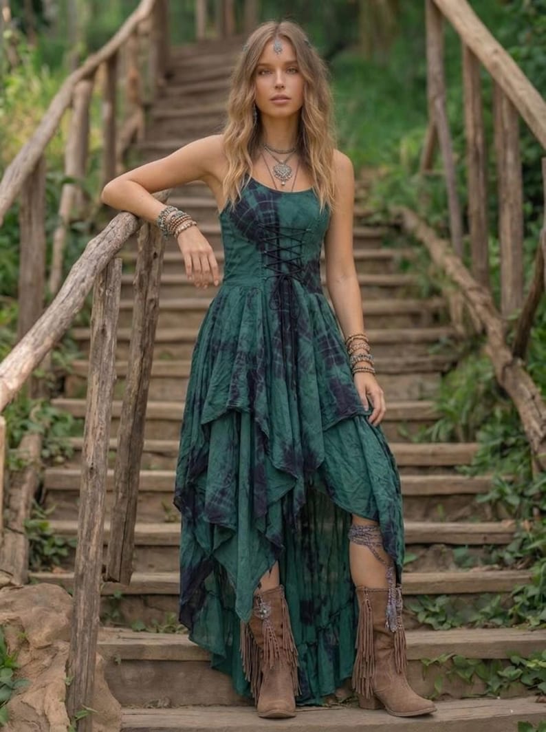 A woman stands on a rustic wooden staircase in a forest, wearing a forest green and black tie-dye fairy-style dress. The dress features a corset-style lace-up bodice and a high-low handkerchief hemline with layered pointed edges. She is styled with bohemian silver jewelry and fringe boots. The lighting is soft and natural, emphasizing the earthy, mystical, and witchy aesthetic of the outfit. Perfect for festivals, woodland ceremonies, or gothic boho fashion