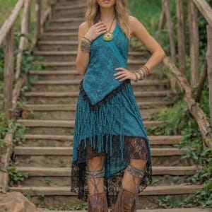 Könnte beinhalten: Ein türkisfarbenes Neckholder-Kleid mit gemustertem Oberteil und Fransenrock, akzentuiert mit schwarzem Spitzensaum. Das Outfit wird mit braunen Fransenstiefeln und mehreren Armbändern kombiniert. Das Model steht auf Holztreppen.