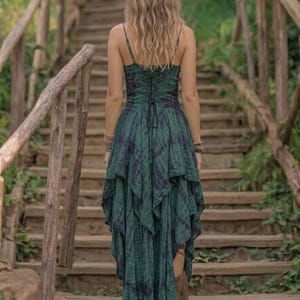 A woman stands on a rustic wooden staircase in a forest, wearing a forest green and black tie-dye fairy-style dress. The dress features a corset-style lace-up bodice and a high-low handkerchief hemline with layered pointed edges. She is styled with bohemian silver jewelry and fringe boots. The lighting is soft and natural, emphasizing the earthy, mystical, and witchy aesthetic of the outfit. Perfect for festivals, woodland ceremonies, or gothic boho fashion