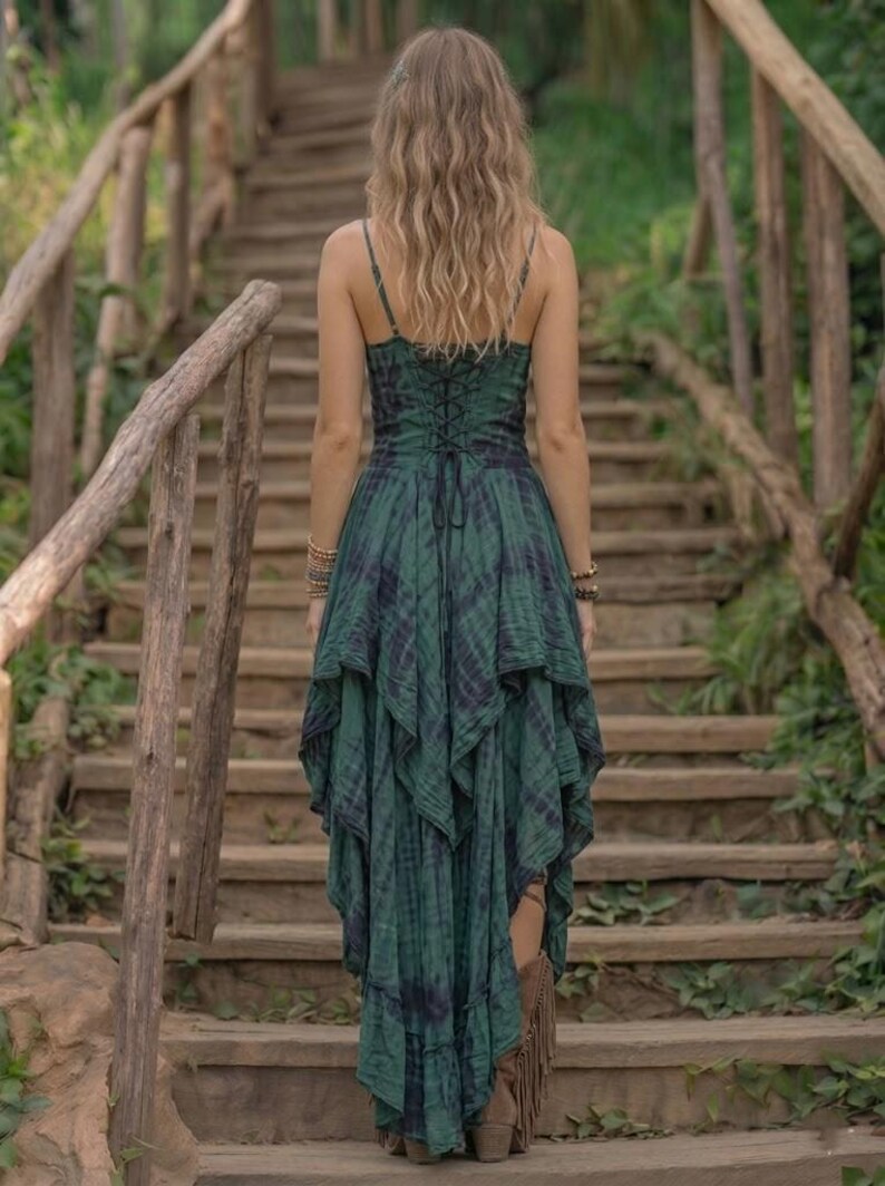 A woman stands on a rustic wooden staircase in a forest, wearing a forest green and black tie-dye fairy-style dress. The dress features a corset-style lace-up bodice and a high-low handkerchief hemline with layered pointed edges. She is styled with bohemian silver jewelry and fringe boots. The lighting is soft and natural, emphasizing the earthy, mystical, and witchy aesthetic of the outfit. Perfect for festivals, woodland ceremonies, or gothic boho fashion