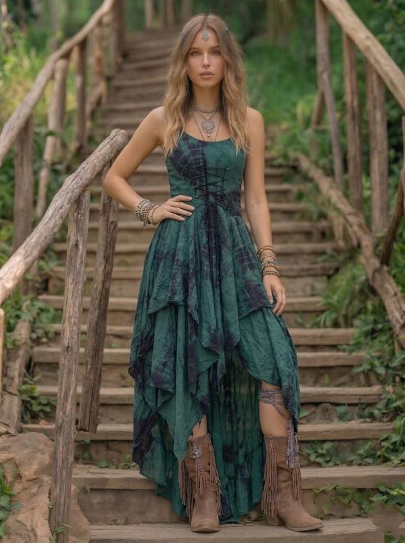 A woman stands on a rustic wooden staircase in a forest, wearing a forest green and black tie-dye fairy-style dress. The dress features a corset-style lace-up bodice and a high-low handkerchief hemline with layered pointed edges. She is styled with bohemian silver jewelry and fringe boots. The lighting is soft and natural, emphasizing the earthy, mystical, and witchy aesthetic of the outfit. Perfect for festivals, woodland ceremonies, or gothic boho fashion