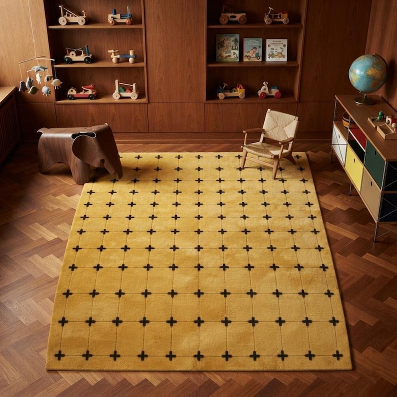 May include: A rectangular area rug featuring a mustard yellow base with a repeating grid pattern of thin black lines and small black cross symbols. The rug is positioned on a wooden floor in a room with wooden shelving units and furniture.