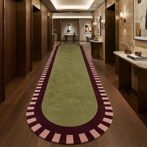 Puede incluir: Una alfombra de pasillo larga y ovalada con un centro de color verde oliva s&oacute;lido. La alfombra est&aacute; bordeada por una banda ancha de color burdeos oscuro, acentuada con un patr&oacute;n repetitivo de bloques rectangulares rosa claro.