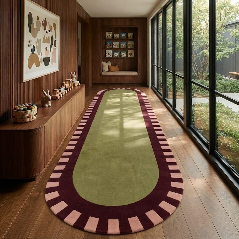 Puede incluir: Una alfombra ovalada alargada con un centro de color verde salvia s&oacute;lido. La alfombra est&aacute; bordeada por una banda ancha de color granate oscuro, acentuada por un patr&oacute;n repetitivo de bloques rectangulares rosa claro en el borde.