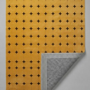 May include: A rectangular area rug featuring a mustard yellow surface with a repeating grid pattern of small black cross symbols. The bottom right corner is folded over to reveal a gray and white herringbone patterned backing material.