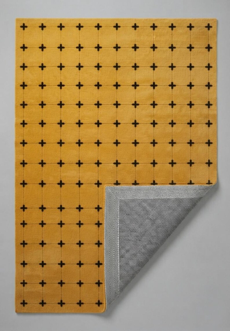 May include: A rectangular area rug featuring a mustard yellow surface with a repeating grid pattern of small black cross symbols. The bottom right corner is folded over to reveal a gray and white herringbone patterned backing material.