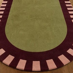 Puede incluir: Una alfombra de forma ovalada con un centro de color verde oliva s&oacute;lido. El borde consiste en una banda de color burdeos oscuro acentuada con un patr&oacute;n repetitivo de bloques rectangulares de color rosa claro en un dise&ntilde;o radial.