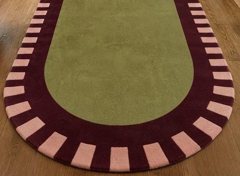 Puede incluir: Una alfombra de forma ovalada con un centro de color verde oliva s&oacute;lido. El borde consiste en una banda de color burdeos oscuro acentuada con un patr&oacute;n repetitivo de bloques rectangulares de color rosa claro en un dise&ntilde;o radial.