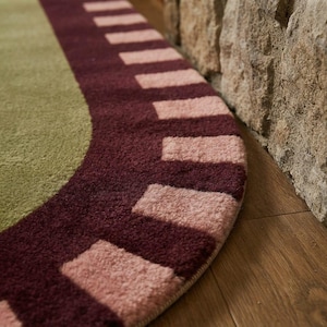 Puede incluir: Una alfombra circular con un centro de color verde salvia s&oacute;lido rodeado por un borde ancho de color burdeos oscuro. El borde est&aacute; decorado con un patr&oacute;n repetitivo de bloques rectangulares de color rosa claro en formaci&oacute;n radial.