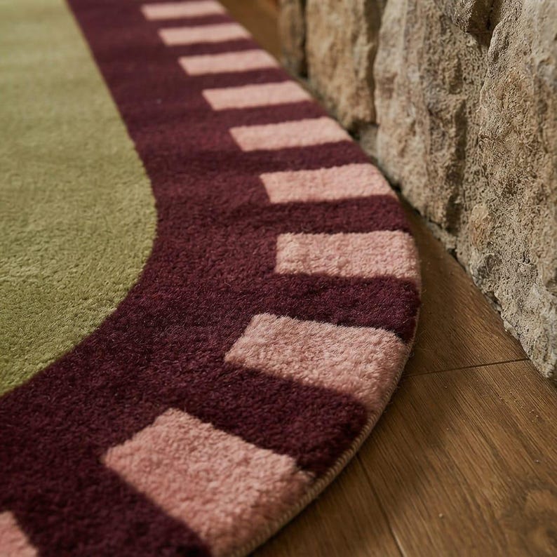 Puede incluir: Una alfombra circular con un centro de color verde salvia s&oacute;lido rodeado por un borde ancho de color burdeos oscuro. El borde est&aacute; decorado con un patr&oacute;n repetitivo de bloques rectangulares de color rosa claro en formaci&oacute;n radial.