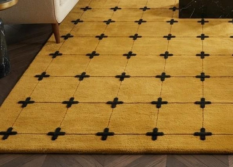 May include: A rectangular area rug featuring a solid mustard yellow background with a grid pattern formed by thin black lines. At each intersection of the grid lines, there is a small, thick black cross shape embroidered into the rug surface.
