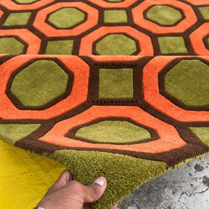 May include: A tufted area rug featuring a repeating geometric pattern of orange octagons and olive green squares outlined in dark brown. The text REGALRUGHOME is printed in black on the surface of the rug, which has a thick, textured pile.