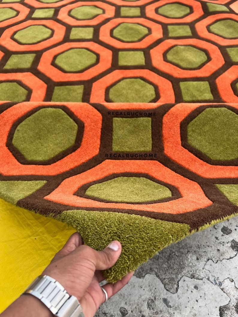 May include: A tufted area rug featuring a repeating geometric pattern of orange octagons and olive green squares outlined in dark brown. The text REGALRUGHOME is printed in black on the surface of the rug, which has a thick, textured pile.