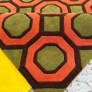 May include: A tufted area rug featuring a repeating geometric pattern of olive green octagons and squares outlined in dark brown, with vibrant orange borders. The text REGALRUGHOME is printed in small black letters on the rug surface.