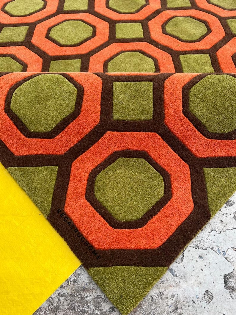 May include: A tufted area rug featuring a repeating geometric pattern of olive green octagons and squares outlined in dark brown, with vibrant orange borders. The text REGALRUGHOME is printed in small black letters on the rug surface.