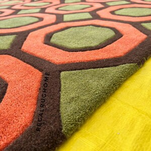 May include: A tufted rug featuring a repeating geometric pattern of orange octagons and green hexagons outlined in dark brown. The text REGALRUGHOME is embossed into the dark brown border of the rug, which has a thick, textured pile surface.