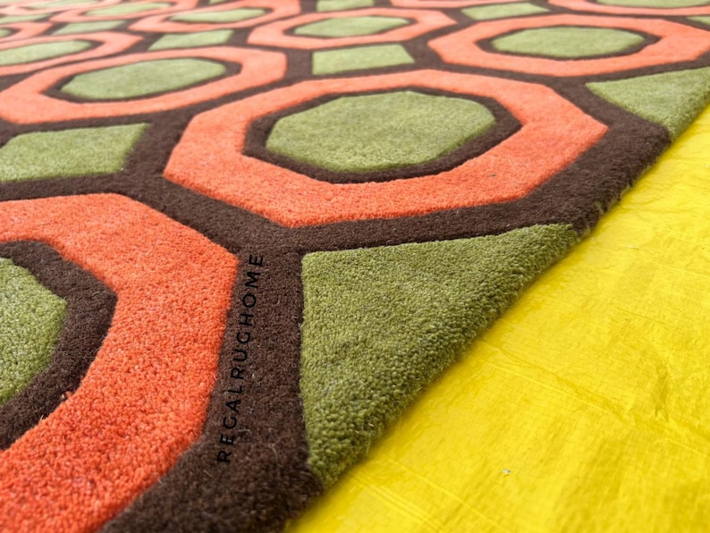 May include: A tufted rug featuring a repeating geometric pattern of orange octagons and green hexagons outlined in dark brown. The text REGALRUGHOME is embossed into the dark brown border of the rug, which has a thick, textured pile surface.