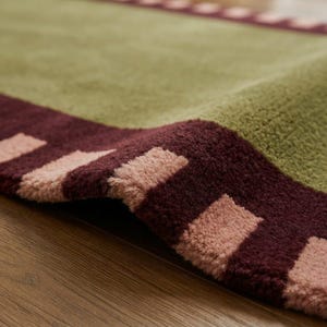 Puede incluir: Una alfombra de felpa con un gran campo central de color verde oliva bordeado por una banda de color burdeos intenso. El borde est&aacute; decorado con un patr&oacute;n repetitivo de bloques rectangulares de color rosa claro sobre la base roja.