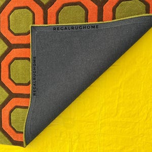 May include: A rug featuring a geometric pattern of orange and dark brown octagons on a lime green background. The rug is folded back to reveal a dark gray backing with the text REGALRUGHOME printed along the edges of the fabric border.
