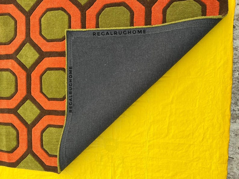 May include: A rug featuring a geometric pattern of orange and dark brown octagons on a lime green background. The rug is folded back to reveal a dark gray backing with the text REGALRUGHOME printed along the edges of the fabric border.