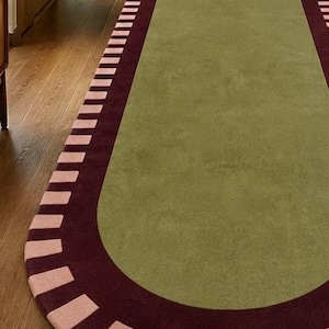 Puede incluir: Una alfombra de pasillo ovalada y alargada con un panel central de color verde oliva s&oacute;lido. La alfombra est&aacute; bordeada por una banda ancha de color burdeos oscuro, acentuada con un patr&oacute;n repetitivo de bloques rectangulares rosa claro.