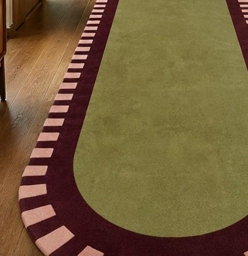 Puede incluir: Una alfombra de pasillo ovalada y alargada con un panel central de color verde oliva s&oacute;lido. La alfombra est&aacute; bordeada por una banda ancha de color burdeos oscuro, acentuada con un patr&oacute;n repetitivo de bloques rectangulares rosa claro.
