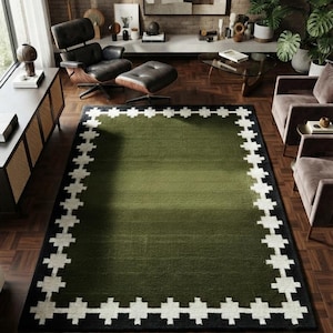 Puede incluir: Una alfombra rectangular con un campo central de color verde oliva sólido. La alfombra está enmarcada por un borde negro grueso que contiene un patrón geométrico repetitivo de cruces blancas a lo largo de los cuatro lados de la pieza.