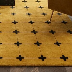 May include: A mustard yellow area rug featuring a grid pattern formed by thin, dark lines. At each intersection of the grid, there is a small, dark, cross-shaped accent. The rug has a soft, plush texture and covers a portion of the floor.