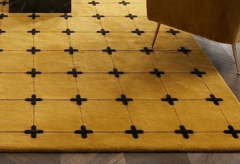 May include: A mustard yellow area rug featuring a grid pattern formed by thin, dark lines. At each intersection of the grid, there is a small, dark, cross-shaped accent. The rug has a soft, plush texture and covers a portion of the floor.