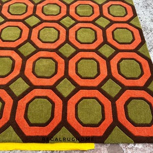 May include: A rectangular area rug featuring a repeating geometric pattern of orange octagons outlined in dark brown, set against a textured olive green background. The text REGALRUGHOME is printed in small black letters near the bottom edge.