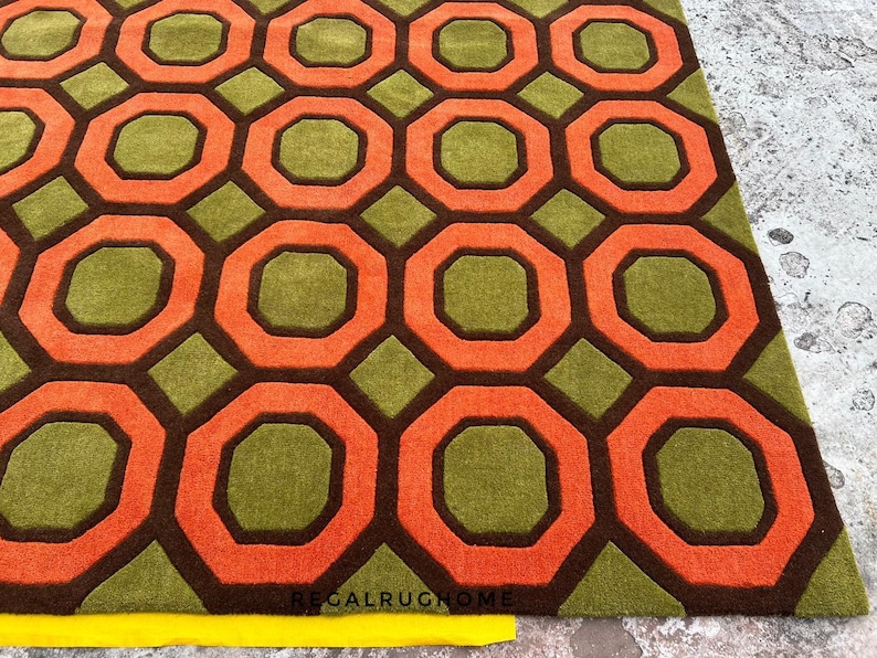 May include: A rectangular area rug featuring a repeating geometric pattern of orange octagons outlined in dark brown, set against a textured olive green background. The text REGALRUGHOME is printed in small black letters near the bottom edge.