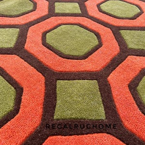 May include: A textured area rug featuring a repeating geometric pattern of olive green octagons and squares outlined in dark brown, set against a vibrant orange background. The text REGALRUGHOME is printed in small black letters near the bottom center.
