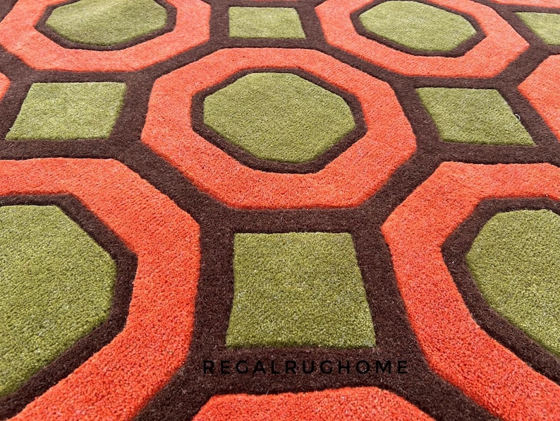 May include: A textured area rug featuring a repeating geometric pattern of olive green octagons and squares outlined in dark brown, set against a vibrant orange background. The text REGALRUGHOME is printed in small black letters near the bottom center.