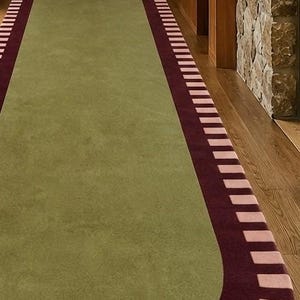 Puede incluir: Una alfombra de pasillo larga con un panel central de color verde oliva s&oacute;lido. La alfombra est&aacute; enmarcada por un borde de color burdeos oscuro acentuado con un patr&oacute;n repetitivo de bloques rectangulares de color rosa claro.