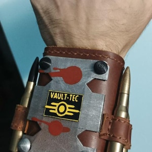 Puede incluir: Una muñequera de cuero marrón con una placa metálica central que muestra el logotipo de Vault-Tec en amarillo y negro. La placa tiene detalles circulares rojos y está asegurada con pernos. Dos casquillos de bala color latón están sujetos a los lados.