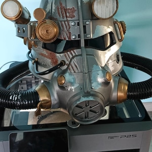 Puede incluir: Un casco de armadura asistida plateado y azul desgastado con accesorios laterales circulares dorados, un aparato respiratorio central y mangueras negras corrugadas. El casco muestra óxido simulado y daños de batalla, sobre una impresora Bambu Lab P2S.