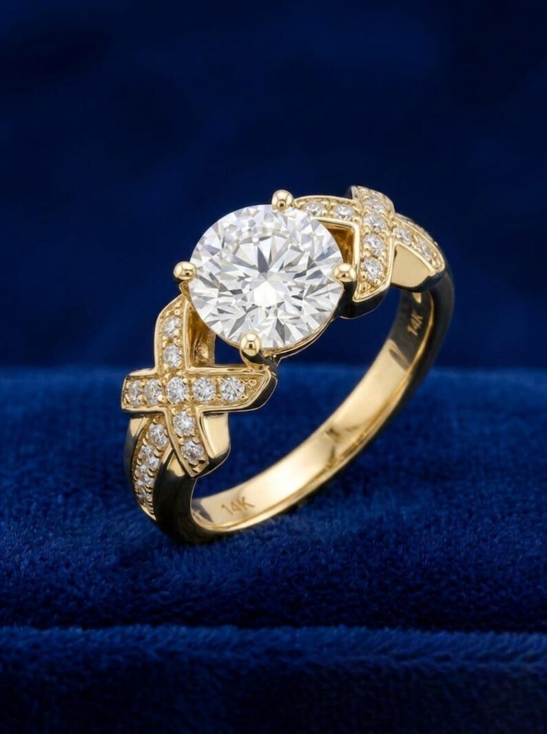 Puede incluir: Un anillo de oro amarillo de 14k con una gran piedra central redonda y transparente de talla brillante. La banda presenta dos motivos en forma de X a cada lado, incrustados con peque&ntilde;as piedras blancas brillantes. El interior de la banda tiene grabado claramente el sello de 14k.