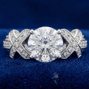 Puede incluir: Un anillo de tono plateado con una gran piedra central redonda y transparente de talla brillante, sujeta por cuatro garras. La banda est&aacute; decorada con dos motivos en forma de X a cada lado, incrustados con peque&ntilde;as piedras transparentes.