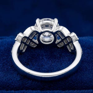 Puede incluir: Un anillo de tono plateado con una gran piedra central redonda y facetada sujeta por un engaste de m&uacute;ltiples garras. La banda presenta un dise&ntilde;o calado intrincado en ambos lados, incorporando piedras claras de corte rectangular.