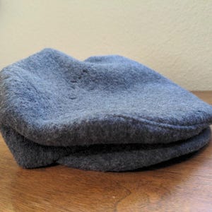 May include: A gray flat cap made of a soft, textured material. The hat is folded and resting on a wooden surface. The cap has a classic, casual style.