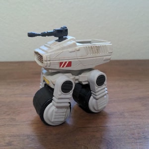 May include: A tan and black toy vehicle with a rotating gun on top. The toy has two large wheels and a rectangular body with red and black accents. The toy is sitting on a wooden surface.