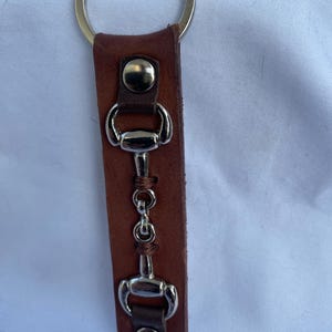 May include: A brown leather keyring featuring a silver-toned metal horse bit ornament secured with two silver-coloured metal snaps. The leather strap is attached to a circular silver-toned metal split ring at the top for keys.