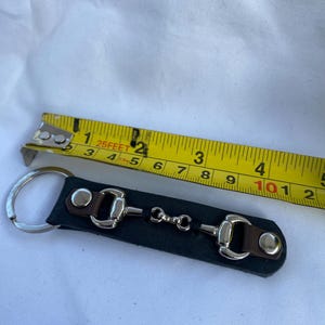 May include: A black leather keyring featuring a silver-toned metal horse bit snaffle detail, brown leather accents, and silver studs. A yellow measuring tape is positioned above the keyring, showing a length of approximately 10.8 centimetres.